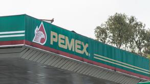 Angled view of Pemex (Petroleos Mexicanos) gas station canopy featuring bold branding, green color scheme, and trees in the background on a cloudy day