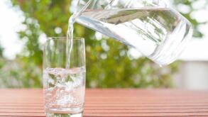 Drinking water flowing from a glass jug into a glass ? background with plants