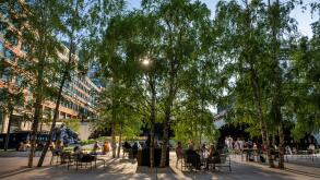 London, UK: Exchange Square at Broadgate, an open space in the City of London. People sit in the square and evening sunlight filters through the trees