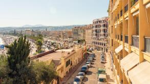 Sunny day view of a European city street lined with colorful buildings, parked cars, and a glimpse of the waterfront in the distance, Nice, FRance
