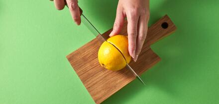 A woman's hands cut a yellow ripe lemon in half with knife on a wooden board on a green background. Top view.