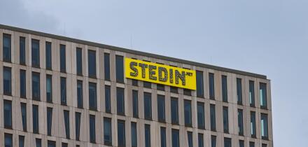 The top corner of the modern Stedin office building, showing its distinctive facade and the company's bright yellow logo against a grey sky. Rotterdam