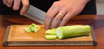 A male hand slicing a cucumber on a wooden cutting board.