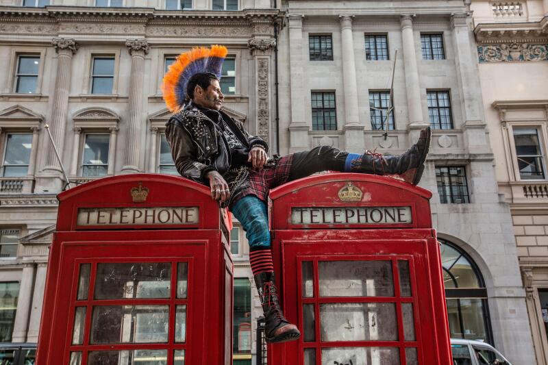 Punk rocker with mohican hair sits relaxing on top of red telephone boxes in central London, England, United Kingdom