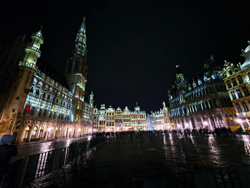 Night view of the illuminated Grand Place in Brussels, Belgium, with historic buildings and crowds gathered in the square.