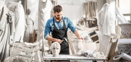 Portrait of a handsome sculptor in blue t-shirt and apron working with stone sculptures on the table at the old atmospheric studio