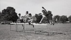 Secondary school sports day, England, c1960s, track & field, four schoolboys competing outside on.a grass track in a hurdles race, leaping over the barriers.