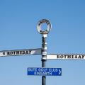 An island road sign on the Isle of Bute, Scotland against a blue sky. It shows the same destination, Rothesay, in two directions.
