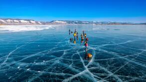 Aerial top view to tourist walk go on smooth surface. People with sleds moves along the blue clear ice. Winter crossing of Lake Baikal. Cracks glossy