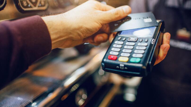 One hand hold machine for cards white another hold dark card for paying money. Men stand at coffee machine