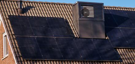 A modern, energy-efficient house in Assen, Netherlands, featuring integrated heat pumps on the roof, solar panels, and a clear blue sky.