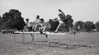 Secondary school sports day, England, c1960s, track & field, four schoolboys competing outside on.a grass track in a hurdles race, leaping over the barriers.