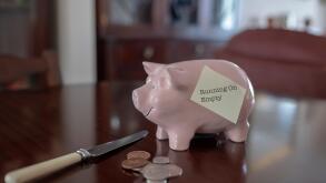 Piggy bank on polished table with loose change and knife and post it note saying running on empty