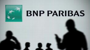 The BNP Paribas logo is seen on an LED screen in the background while a silhouetted person uses a smartphone in the foreground (Editorial use only)