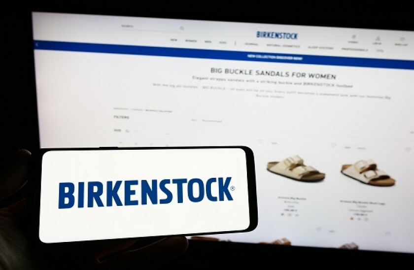 Person holding smartphone with logo of shoe company Birkenstock Group B.V. Co. KG on screen in front of website. Focus on phone display.